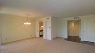 This single-floor space presents an open-concept living and dining area finished with neutral-toned carpet and white walls, creating a bright and understated atmosphere. An elegant chandelier anchors the dining area, adding a classic touch to the otherwise simple aesthetic. The layout flows naturally, with a partial wall separating the main living space from the kitchen beyond. The kitchen features warm wood cabinetry, a dark countertop, and a dishwasher, accessible through a doorway that maintains a sense of openness while defining the cooking area. A second doorway visible from the main room leads to additional areas of the unit, suggesting a practical and well-organized floor plan. HVAC vents are integrated into the ceiling and walls, ensuring year-round comfort. The consistent use of light, neutral tones throughout — from the carpet to the walls and ceiling — contributes to a cohesive and airy feel across the space. Overall, the layout is straightforward and functional, suited to everyday living.