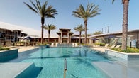 This outdoor pool area is designed to offer a full resort-style experience. The centerpiece is a generously sized swimming pool with clear, blue water, accompanied by a separate spa. The pool deck provides ample space for lounge chairs and umbrella-shaded seating, making it well-suited for relaxation and recreation.

Towering palm trees frame the primary pool area, adding a tropical aesthetic and providing natural shade throughout the day. The surrounding landscaping is thoughtfully maintained, contributing to an inviting, resort-like ambiance. The pool house structure nearby features a multi-level architectural design with warm earth tones and serves as an attractive visual backdrop to the space.

A secondary pool area is also present, featuring a classic rectangular pool layout surrounded by a wide concrete deck lined with lounge chairs and patio umbrellas. A pool house with dark wood-framed doors and stucco exterior walls provides access to this area, with large glass sliding doors offering a direct view of the pool from inside. The surrounding multi-story buildings with brick and wood siding provide a defined, community-oriented setting. Together, both pool areas deliver a well-rounded outdoor amenity space suitable for leisure and social gatherings.