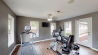 Located at 2144 Lakeshore Dr in Ridgeland, Mississippi, this single-floor fitness center is a well-equipped space designed to support a range of cardio-focused workouts. The room is finished with warm hardwood-style flooring that contrasts neatly against the cool gray walls and crisp white baseboards, creating a clean and functional aesthetic throughout.

The space is outfitted with a comprehensive selection of cardio equipment, including a treadmill, elliptical trainer, upright stationary bike, and a recumbent exercise bike, offering users a variety of workout options. A wall-mounted flat-screen television is positioned for easy viewing during exercise, and a ceiling fan with a light fixture provides both air circulation and ambient lighting.

Multiple windows line the walls, allowing generous natural light to fill the room while offering views of the surrounding outdoor greenery. A water cooler station is conveniently available, ensuring hydration is within easy reach. The space also features a posted fitness rules or cardio notes board, indicating an organized and community-oriented environment. Access to the outdoors is provided through a glass-panel door, further enhancing the bright and open feel of the facility.