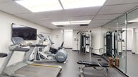 Located at 3920 N 56th Ave in Hollywood, Florida, this fitness center is a single-floor facility designed to support a comprehensive workout experience. The space is organized into distinct functional zones, accommodating both cardiovascular training and strength work within a practical, efficiently arranged layout.

The cardio area is equipped with multiple Precor and Matrix treadmills, elliptical trainers, and a recumbent stationary bike, providing a range of low- and high-impact exercise options. A wall-mounted flat-screen television is positioned to be visible from the cardio equipment, enhancing the workout experience. Natural light filters in through windows fitted with roller shades, offering views of the surrounding outdoor greenery.

The strength training area features a multi-station cable machine suitable for a variety of resistance exercises, an adjustable weight bench, and a well-stocked dumbbell rack. Full-length mirrored panels line one wall, visually expanding the space while allowing users to monitor their form. The rubber flooring throughout provides durability and safety underfoot.

The drop ceiling with recessed panel lighting ensures consistent, even illumination across the entire facility. A hand sanitizer dispenser and paper towel station indicate attention to hygiene. Informational postings are displayed near the entrance area, completing the functional, user-focused environment.