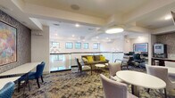This single-floor amenity space offers a refined and well-appointed environment designed for comfort and functionality. The central lounge area is the heart of the floor, featuring a mix of plush seating including a rich olive-toned sofa accented with vibrant yellow throw pillows, upholstered armchairs, and a variety of round tables suited for casual gatherings or individual use. Elegant petal-shaped pendant light fixtures hang from a coffered white ceiling, casting warm, inviting light throughout. A glass railing runs along the lounge's perimeter, allowing natural light to filter in through a row of clerestory windows that frame views of surrounding greenery. A dedicated work or study nook features bar-height tables and cobalt blue chairs set against a dramatic textured wallcovering, anchored by a large, colorful abstract painting. Adjacent to this area, a kitchenette is outfitted with dark wood cabinetry, granite countertops, a stainless steel sink, and modern appliances including an ice and water dispenser. A fitness center, visible through large glass-paned doors, houses cycling equipment and mirrors, offering a bright and motivating workout environment. Corridors connecting the various areas are thoughtfully finished with the same textured wallcovering and patterned carpeting, with framed artwork adding artistic continuity throughout.