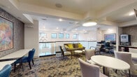 This single-floor amenity space offers a refined and well-appointed environment designed for comfort and functionality. The central lounge area is the heart of the floor, featuring a mix of plush seating including a rich olive-toned sofa accented with vibrant yellow throw pillows, upholstered armchairs, and a variety of round tables suited for casual gatherings or individual use. Elegant petal-shaped pendant light fixtures hang from a coffered white ceiling, casting warm, inviting light throughout. A glass railing runs along the lounge's perimeter, allowing natural light to filter in through a row of clerestory windows that frame views of surrounding greenery. A dedicated work or study nook features bar-height tables and cobalt blue chairs set against a dramatic textured wallcovering, anchored by a large, colorful abstract painting. Adjacent to this area, a kitchenette is outfitted with dark wood cabinetry, granite countertops, a stainless steel sink, and modern appliances including an ice and water dispenser. A fitness center, visible through large glass-paned doors, houses cycling equipment and mirrors, offering a bright and motivating workout environment. Corridors connecting the various areas are thoughtfully finished with the same textured wallcovering and patterned carpeting, with framed artwork adding artistic continuity throughout.
