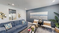 Located at 222 Kingfisher Way in Royal Palm Beach, Florida, this thoughtfully designed single-floor space delivers a polished coastal-contemporary aesthetic. The interior is anchored by a bold slate-blue textured accent wall that adds depth and visual interest throughout the main living area. Light-toned wood-style flooring runs seamlessly underfoot, complementing the recessed lighting and white trim that brighten the space.

The main lounge area is open and well-appointed, featuring generous seating, a sleek coffee table, and a large-scale landscape artwork that reinforces the coastal theme. A light wood media console houses a wall-mounted flat-screen TV, flanked by windows with white blinds that allow natural light to filter in.

A dedicated wet bar area features navy-blue cabinetry with white countertops, a sink, and a compact refrigerator — ideal for entertaining or refreshment needs. Decorative touches such as a sunburst mirror, geometric wall accents, and curated greenery elevate the ambiance throughout.

The entrance provides a clean, organized transition into the space, while the balcony extends the experience outdoors. Every detail, from the carefully selected furnishings to the cohesive color palette, contributes to a warm, functional, and visually refined environment.