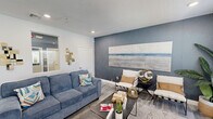 Located at 222 Kingfisher Way in Royal Palm Beach, Florida, this thoughtfully designed single-floor space delivers a polished coastal-contemporary aesthetic. The interior is anchored by a bold slate-blue textured accent wall that adds depth and visual interest throughout the main living area. Light-toned wood-style flooring runs seamlessly underfoot, complementing the recessed lighting and white trim that brighten the space.

The main lounge area is open and well-appointed, featuring generous seating, a sleek coffee table, and a large-scale landscape artwork that reinforces the coastal theme. A light wood media console houses a wall-mounted flat-screen TV, flanked by windows with white blinds that allow natural light to filter in.

A dedicated wet bar area features navy-blue cabinetry with white countertops, a sink, and a compact refrigerator — ideal for entertaining or refreshment needs. Decorative touches such as a sunburst mirror, geometric wall accents, and curated greenery elevate the ambiance throughout.

The entrance provides a clean, organized transition into the space, while the balcony extends the experience outdoors. Every detail, from the carefully selected furnishings to the cohesive color palette, contributes to a warm, functional, and visually refined environment.