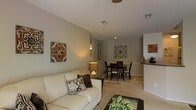 This single-floor residence offers a thoughtfully designed open-concept layout that connects the living, dining, and kitchen areas into one cohesive and functional space. The living area is anchored by a plush sofa set against warm, neutral-toned walls, complemented by a glass-top coffee table and decorative wall art that adds character to the room. Soft, wall-to-wall carpeting runs throughout, contributing to a cozy and comfortable ambiance. Adjacent to the living area, the dining space accommodates a dark wood table and chair set, highlighted by recessed lighting overhead and tasteful wall décor. The layout allows for a natural flow between spaces, making the area well-suited for both everyday living and entertaining. A glimpse into the kitchen reveals dark countertops and a clean, efficient design that integrates seamlessly with the open floor plan. Ceiling-mounted ventilation and recessed lighting are distributed throughout, ensuring consistent comfort and illumination. The textured ceiling adds a subtle architectural detail, while the neutral color palette creates a bright and welcoming atmosphere throughout the home.