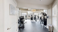 Located at 7880 Triangle Promenade Dr in Raleigh, NC, this fitness center offers a thoughtfully designed exercise space that combines functionality with an airy, welcoming atmosphere. The single-floor layout provides a generous open floor plan, making it easy to navigate between equipment stations.

The exercise room is defined by soaring ceilings and expansive floor-to-ceiling windows that line the walls, flooding the space with natural light and offering views of the surrounding wooded landscape. Large drum pendant light fixtures and ceiling fans add to the polished, well-appointed aesthetic while ensuring comfortable air circulation throughout.

The room is outfitted with a comprehensive selection of strength training machines and cardiovascular equipment, including treadmills, ellipticals, and stationary bikes — all featuring integrated display screens. Full-length wall mirrors on one side of the room visually expand the space and assist users in monitoring their form during workouts.

Durable rubber flooring in a neutral gray tone covers the entire exercise area, providing both safety and easy maintenance. The clean, neutral color palette of white walls and warm beige tones contributes to a bright and motivating environment. A posted fitness center rules board is visible near the entrance, reflecting an organized and well-managed facility.