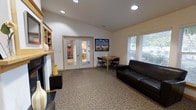 This single-floor library offers a warm and functional environment designed for reading, study, and quiet gathering. The main room features carpeted flooring and a clean, open layout that promotes a comfortable and inviting atmosphere. A decorative fireplace with a wooden mantle and built-in shelving anchors one side of the room, adding a classic, homey touch to the space.

A dark leather sofa is positioned near a window fitted with slatted blinds, allowing natural light to filter into the room while maintaining privacy. Adjacent to the window, a small wooden table with folding chairs provides a casual workspace or study area. Colorful artwork displayed on the wall adds visual interest and character to the otherwise neutral, white-walled interior.

Recessed ceiling lighting ensures the space remains well-lit throughout, complementing the natural light from the windows. Glass-paneled double doors lead to an adjoining room visible in the background, which appears to feature additional work or activity areas, suggesting the library offers multiple functional zones. The overall ambiance is calm, approachable, and purposefully arranged to support a variety of library activities in a comfortable, community-oriented setting.