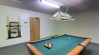 This single-floor recreational space is designed with leisure and social activity in mind. The room's standout feature is a full-size pool table with a green felt surface, set beneath a decorative stained-glass-style pendant light suspended by chains from the ceiling. The pendant fixture adds a classic, inviting ambiance to the room, casting warm, focused light directly over the playing surface. The walls are painted in a soft, neutral tone, contributing to a clean and open atmosphere, while the textured ceiling adds a subtle layer of character. Practical fluorescent lighting supplements the pendant light to ensure the room is well-illuminated throughout. The space features durable, easy-to-maintain flooring with a simple baseboard trim, keeping the aesthetic functional and straightforward. A wooden door provides access to an adjoining area, making this room easily accessible within the broader layout. Overall, this recreational room offers a comfortable and purposeful environment well-suited for casual games and community gatherings.