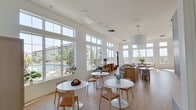 This single-floor community gathering space is defined by its airy, open layout and an abundance of natural light. Soaring ceilings amplify the sense of spaciousness throughout, while expansive floor-to-ceiling windows fitted with transom panels line multiple walls, offering unobstructed views of an outdoor pool area and surrounding landscaping. Light hardwood flooring runs continuously across the entire space, reinforcing the clean, modern aesthetic.

The main area is arranged with multiple round white pedestal tables paired with natural wood and woven-leather chairs, providing flexible seating configurations suited for both casual gatherings and more structured events. A built-in window bench along one wall offers additional seating with direct pool views. A central wood-toned island with counter seating serves as a functional focal point, complemented by a large pendant light overhead.

A dedicated service area along one wall features pale blue cabinetry with ample upper storage and a clean white countertop, ideal for catering or event setup. A wall-mounted television and additional lounge seating are positioned to take advantage of the natural light streaming through the tall windows. Direct outdoor access is provided via a glass-paneled door. The overall ambiance is bright, minimal, and welcoming.