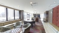 This refined lounge space offers an inviting and upscale atmosphere designed for comfort and social interaction. The defining feature of the room is its expansive floor-to-ceiling windows that wrap around the perimeter, flooding the interior with natural light and providing sweeping panoramic views of the surrounding cityscape and neighboring high-rise buildings. The seating area is thoughtfully curated, featuring tufted armchairs, barrel-back lounge chairs, and small round tables arranged to encourage both intimate conversation and relaxed gatherings. A richly patterned area rug in muted tones of burgundy, taupe, and grey anchors the central seating zone, adding warmth and visual interest to the space. The flooring transitions to light wood-toned planks toward the perimeter, complementing the neutral white ceiling with recessed and flush-mounted lighting fixtures. Toward the rear of the space, a kitchenette or servery area is visible, along with additional seating arrangements and a pendant light, suggesting a multifunctional design suited for events or everyday use. An accent wall with textured stone or tile detail adds an architectural focal point, further elevating the overall aesthetic. This space combines modern design sensibilities with comfortable, hospitality-inspired furnishings.