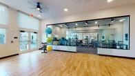 This fitness center is thoughtfully designed to accommodate a full range of exercise activities across two distinct zones connected by an open, airy layout. The open studio area features warm hardwood flooring, high ceilings, and a ceiling fan, creating a bright and inviting environment ideal for stretching, yoga, or group fitness. Large windows and glass doors allow abundant natural light to flow throughout, with roller shades providing adjustable light control. Decorative framed artwork adorns the white walls, adding a polished, welcoming atmosphere. A collection of exercise balls and a stationary bike round out the studio's functional versatility. Separating the studio from the equipment zone is a sleek floor-to-ceiling glass partition with a dark-framed sliding door, offering visual openness while defining each area. The equipment zone is carpeted with durable rubber-backed flooring and houses an extensive array of strength training machines, including cable machines, seated press stations, and various resistance equipment. A large wall mirror enhances the sense of space within this section. An adjoining cardio room features a row of treadmills and stationary bikes positioned near windows for natural light, completing a comprehensive and well-appointed fitness facility.