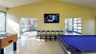 This single-floor recreation room is a well-appointed communal space designed for leisure and entertainment. The main game area is generously sized, featuring polished concrete floors that reflect natural light streaming through large glass doors and windows. Two blue-felt pool tables serve as centerpieces, complemented by a foosball table. A row of seating chairs faces a wall-mounted flat-screen TV beneath a vaulted yellow-painted ceiling, creating a comfortable viewing area. Adjacent fitness alcoves house cable machines and a treadmill, accessible through open pass-through openings that maintain visual connectivity throughout the space. Bold burgundy accent walls contrast with the neutral white tones throughout, adding visual depth. The kitchen area is thoroughly equipped with stainless steel appliances including a gas range, refrigerator, dishwasher, and microwave, all set against gray flat-panel cabinetry and a tile mosaic backsplash. White countertops extend to a peninsula with bar seating. A window above the sink brings in natural light and outdoor views. Cue racks, decorative wall art, and ample open floor space complete this versatile and functional recreation facility.