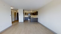 Located at 131 Rantoul in Beverly, Massachusetts, this modern single-floor residence offers a clean, contemporary design with an open and airy feel. The space features light-toned hardwood flooring that extends throughout, lending warmth and continuity to the layout.

The kitchen is a standout feature, designed with dark two-tone cabinetry — rich upper cabinets paired with sleek lower grey units — and a light-colored countertop that provides strong visual contrast. Stainless steel appliances, including a range, microwave, and refrigerator, are seamlessly integrated into the design. A peninsula-style counter extends into the living area, creating a natural divide between the kitchen and the open living space while also offering additional prep and seating space.

Track lighting overhead illuminates the kitchen efficiently, while recessed lighting contributes to the overall brightness of the space. The walls are finished in a crisp, neutral white, enhancing the sense of openness. A hallway visible in the background suggests additional rooms beyond the main living area.

The open-concept design connects the kitchen and living areas effortlessly, making the space feel expansive and well-suited for both everyday living and entertaining. The overall aesthetic is modern, minimal, and well-appointed.