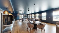 Located at 808 Melvin Ct in Madison, WI, this single-floor community lounge offers a well-appointed, thoughtfully designed gathering space. The interior is defined by a cohesive color palette of deep navy and steel blue walls paired with warm light wood flooring, creating a sophisticated yet inviting atmosphere throughout.

The main living area serves as the social hub, featuring multiple round tables with upholstered chairs arranged on textured area rugs, providing flexible seating for casual gatherings or conversation. A dedicated lounge zone offers comfortable armchairs, a wall-mounted TV, and large windows that flood the space with natural light and provide views to the outdoors.

Along one wall, a striking geometric-patterned accent wall in blue and white sets the backdrop for a row of intimate banquette booths with plush cushioned seating and warm amber pendant lighting overhead — ideal for smaller group settings. A service bar area with decorative pendant lights and tiled backsplash anchors one end of the space.

A private meeting or conference nook features distinctive patterned wallpaper, soft mint-green wainscoting, a round table, and leather seating, offering a quieter retreat within the larger layout. The deep blue ceiling treatment adds depth and visual continuity across all areas, tying the design together with a polished, modern aesthetic.
