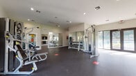 This fitness center is a thoughtfully designed, single-floor facility that offers a comprehensive workout environment. The space is organized to accommodate a wide range of exercise activities, from cardiovascular training to strength and resistance work.

The cardio zone is lined with TRUE brand treadmills and elliptical machines, positioned alongside a row of large windows that frame views of the surrounding lush, tropical greenery. Wall-mounted flat-screen televisions are installed above the cardio equipment, providing entertainment during workouts. A water station is conveniently located nearby.

The strength training area features a multi-functional cable machine, a dumbbell rack, and additional resistance equipment. Full-length mirrors are strategically placed along multiple walls, enhancing the sense of openness and allowing users to monitor their form throughout their workouts.

The facility is finished with durable dark rubber flooring throughout, providing both safety and comfort underfoot. Recessed lighting and a textured ceiling contribute to a clean, functional aesthetic. Large glass doors along one wall open to views of the outdoor surroundings, including palm trees, flooding the space with natural light and creating an inviting, airy atmosphere. The overall design balances functionality with a modern, well-maintained aesthetic suitable for a broad range of fitness needs.