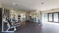 This fitness center is a thoughtfully designed, single-floor facility that offers a comprehensive workout environment. The space is organized to accommodate a wide range of exercise activities, from cardiovascular training to strength and resistance work.

The cardio zone is lined with TRUE brand treadmills and elliptical machines, positioned alongside a row of large windows that frame views of the surrounding lush, tropical greenery. Wall-mounted flat-screen televisions are installed above the cardio equipment, providing entertainment during workouts. A water station is conveniently located nearby.

The strength training area features a multi-functional cable machine, a dumbbell rack, and additional resistance equipment. Full-length mirrors are strategically placed along multiple walls, enhancing the sense of openness and allowing users to monitor their form throughout their workouts.

The facility is finished with durable dark rubber flooring throughout, providing both safety and comfort underfoot. Recessed lighting and a textured ceiling contribute to a clean, functional aesthetic. Large glass doors along one wall open to views of the outdoor surroundings, including palm trees, flooding the space with natural light and creating an inviting, airy atmosphere. The overall design balances functionality with a modern, well-maintained aesthetic suitable for a broad range of fitness needs.