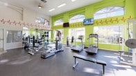 This fitness center is designed to energize and motivate with its bold, vibrant aesthetic and comprehensive equipment offerings. The space is defined by striking lime-green accent walls adorned with a red heartbeat graphic, creating a dynamic and health-focused atmosphere. Large arched windows line the walls, flooding the room with abundant natural light and offering views of the surrounding greenery outdoors.

The facility is well-appointed with a diverse selection of cardio equipment, including multiple treadmills and an elliptical machine, catering to a range of workout preferences. A dedicated strength training area features weight machines and free weight stations, supported by floor-to-ceiling mirrors that allow users to monitor their form. Flat-screen televisions are mounted on the walls, providing entertainment during workouts.

The flooring consists of durable, dark rubberized material suited for high-impact activity, while the high ceilings contribute to a spacious and open feel. A ceiling fan enhances air circulation throughout the space. Overall, the fitness center combines functional design with an energetic visual identity, offering a well-rounded environment for both cardio and strength-based training routines.