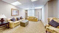 This single-floor suite offers a generously sized, welcoming environment designed for comfort and functionality. The main lounge area is expansive and open, featuring a drop-panel ceiling with recessed lighting panels that provide bright, even illumination throughout. Patterned carpeting in soft, neutral tones runs throughout the space, contributing to a warm and inviting ambiance. Vertical blinds on multiple windows allow for adjustable natural light, connecting the interior to exterior views.

The main lounge is furnished with multiple seating arrangements, including striped and upholstered sofas accented with decorative pillows, end tables with classic table lamps, and framed artwork on the walls. A central structural column anchors the open layout, while a dedicated seating alcove provides a quieter, more intimate space with its own sofa, lamp tables, and a decorative wall mirror.

A separate dining nook accommodates a round glass-top table with metal chairs, positioned beside large windows that admit natural light. The compact kitchenette is efficiently laid out with warm honey-toned wood cabinetry, a full-size refrigerator, a microwave, a sink, and ample counter space, making it well-suited to supporting gatherings or extended stays. Together, these areas form a cohesive, comfortable suite ideal for communal use or hospitality purposes.