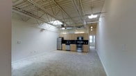 This single-floor loft-style unit showcases an industrial-inspired design defined by its dramatic exposed steel truss ceiling system, which soars overhead and lends the space a sense of openness and architectural character. Ductwork and structural beams are left deliberately visible, complementing the loft aesthetic while a large ceiling fan provides both function and visual continuity within the open volume.

The layout is open-concept, seamlessly integrating the living area and kitchen into one expansive, flowing space. Neutral-toned carpeting covers the living area, offering warmth and contrast against the harder industrial elements above. Natural light filters in through windows visible near the entry corridor, brightening the interior.

The kitchen is well-appointed and functional, featuring light wood cabinetry paired with dark countertops for a clean contrast. A full suite of stainless steel appliances includes a refrigerator, stove, built-in microwave, and dishwasher. The kitchen floor transitions to a hard surface, clearly delineating the cooking area from the carpeted living space.

A hallway visible toward the rear of the unit suggests additional rooms or private spaces beyond the main open area. Overall, the unit combines the raw, open character of industrial loft design with modern finishes and practical amenities.