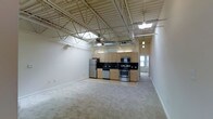 This single-floor loft-style unit showcases an industrial-inspired design defined by its dramatic exposed steel truss ceiling system, which soars overhead and lends the space a sense of openness and architectural character. Ductwork and structural beams are left deliberately visible, complementing the loft aesthetic while a large ceiling fan provides both function and visual continuity within the open volume.

The layout is open-concept, seamlessly integrating the living area and kitchen into one expansive, flowing space. Neutral-toned carpeting covers the living area, offering warmth and contrast against the harder industrial elements above. Natural light filters in through windows visible near the entry corridor, brightening the interior.

The kitchen is well-appointed and functional, featuring light wood cabinetry paired with dark countertops for a clean contrast. A full suite of stainless steel appliances includes a refrigerator, stove, built-in microwave, and dishwasher. The kitchen floor transitions to a hard surface, clearly delineating the cooking area from the carpeted living space.

A hallway visible toward the rear of the unit suggests additional rooms or private spaces beyond the main open area. Overall, the unit combines the raw, open character of industrial loft design with modern finishes and practical amenities.