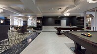 Located at 223 E Broad St in the heart of Richmond, Virginia, this elegantly appointed amenity floor delivers a refined communal experience through a series of thoughtfully designed spaces. The grand main lounge sets the tone with its striking black-and-white patterned tile flooring, coffered ceilings, and bold dark accent shelving with gold trim. Plush wingback and accent chairs in muted tones invite relaxed conversation, while pendant chandeliers cast a warm, sophisticated glow throughout. A dedicated billiards area features multiple pool tables set against dramatic dark patterned wallpaper and wainscoting, creating an atmosphere reminiscent of a classic gentlemen's club with a modern edge. Adjacent to the game area, a casual lounge nook with tufted green velvet seating, decorative wall panels, and a wall-mounted display screen offers a more intimate retreat. The fully equipped kitchen is outfitted with stainless steel appliances, double wall ovens, ample white cabinetry with dark countertops, and open shelving — designed to support events and gatherings of any scale. A separate dining area provides stylish seating around sleek black tables paired with teal and gray velvet chairs, accented by framed artwork and a warm chandelier. Together, these spaces form a cohesive, upscale environment ideal for socializing and entertaining.