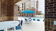 This outdoor amenity space is designed for relaxation and social gathering, centered around a generously sized swimming pool with a smooth, light-toned concrete deck. The pool area is outfitted with multiple lounge chairs and dining sets, each shaded by vibrant blue umbrellas, creating a cohesive and resort-inspired aesthetic throughout. A standout feature of this space is the covered cabana, constructed with rich, dark brick columns and walls that lend a sturdy, classic character to the area. The cabana houses a built-in outdoor kitchen complete with a stainless steel grill and counter space, making it well-suited for outdoor entertaining. High-top dining tables and bar-height seating are positioned within the cabana, offering a comfortable spot to dine al fresco. The deck extends to an open-air lounge zone furnished with deep-seated outdoor sofas, ideal for casual conversation. Mature palm trees are planted throughout the perimeter, adding a tropical feel and softening the fenced boundary of the space. The backdrop is anchored by a multi-story brick and siding residential building with large windows and private balconies, providing a sense of enclosure and community. Overall, the space balances architectural character with practical outdoor amenities.