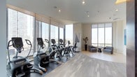This well-appointed fitness center in Dallas is designed to offer an energizing workout environment with a bright, contemporary aesthetic. The space is defined by expansive floor-to-ceiling windows that wrap around the room, flooding the interior with abundant natural light and framing broad views of the surrounding cityscape—creating an open, airy atmosphere that enhances the exercise experience.

The main workout area is outfitted with a row of high-quality cardio machines, including ellipticals and upright bikes, positioned to take full advantage of the panoramic views. Equipment is arranged on protective rubber mats set atop light wood-toned flooring, which adds warmth and visual texture throughout the space.

A dedicated lounge corner provides a comfortable retreat, furnished with modern upholstered chairs and a sleek side table—ideal for cooling down or socializing. Neutral-toned walls and recessed lighting contribute to the clean, polished interior, while tasteful decorative accents, including a potted plant and framed artwork, add subtle character. The overall ambiance is sophisticated, functional, and inviting.