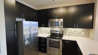 This single-floor space showcases a thoughtfully designed kitchen that serves as a standout feature of the home. The kitchen is outfitted with rich, dark espresso-toned cabinetry that extends from floor to ceiling, maximizing storage while creating a bold, contemporary aesthetic. The cabinetry is complemented by clean white countertops that provide a striking visual contrast, lending the space a crisp and polished look.

A full suite of stainless steel appliances includes a side-by-side refrigerator with an in-door water and ice dispenser, an electric range with a smooth cooktop, and an over-the-range microwave — all contributing to a cohesive, modern finish. A decorative mosaic tile backsplash in neutral tones of white, gray, and brown adds texture and visual interest between the countertops and upper cabinets, serving as an elegant focal point.

The layout of the kitchen is functional and efficient, with well-organized counter space and cabinetry designed to accommodate everyday cooking needs. Sleek bar-pull hardware on the cabinet doors and drawers reinforces the contemporary design language throughout. The overall ambiance of the space is clean, modern, and well-appointed, reflecting a stylish yet practical approach to interior design.