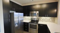 This single-floor space showcases a thoughtfully designed kitchen that serves as a standout feature of the home. The kitchen is outfitted with rich, dark espresso-toned cabinetry that extends from floor to ceiling, maximizing storage while creating a bold, contemporary aesthetic. The cabinetry is complemented by clean white countertops that provide a striking visual contrast, lending the space a crisp and polished look.

A full suite of stainless steel appliances includes a side-by-side refrigerator with an in-door water and ice dispenser, an electric range with a smooth cooktop, and an over-the-range microwave — all contributing to a cohesive, modern finish. A decorative mosaic tile backsplash in neutral tones of white, gray, and brown adds texture and visual interest between the countertops and upper cabinets, serving as an elegant focal point.

The layout of the kitchen is functional and efficient, with well-organized counter space and cabinetry designed to accommodate everyday cooking needs. Sleek bar-pull hardware on the cabinet doors and drawers reinforces the contemporary design language throughout. The overall ambiance of the space is clean, modern, and well-appointed, reflecting a stylish yet practical approach to interior design.