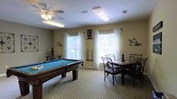 This single-floor recreational space is designed for social activity and leisurely enjoyment. The main room serves as a combined game and gathering area, highlighted by a full-size pool table with a teal felt surface at its center, surrounded by neutral-toned carpeting that adds warmth underfoot. Cue racks and billiard accessories are thoughtfully mounted along the walls, keeping the space organized and ready for play. A ceiling fan overhead provides comfortable air circulation throughout the room. Adjacent to the billiards area, a dining set with dark wooden chairs and a matching table offers a dedicated spot for casual seating or tabletop games. The room is generously illuminated by multiple large windows dressed with white curtains, allowing abundant natural light to pour in and create a bright, welcoming atmosphere. Fluorescent overhead lighting supplements the natural light, ensuring the space remains well-lit at all times. The neutral, warm-toned walls contribute to the relaxed and inviting ambiance of the room. Overall, this recreational space is thoughtfully arranged to accommodate a variety of social activities, making it an ideal venue for community engagement and informal gatherings.