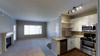 This single-floor space offers a thoughtfully designed open-concept layout that seamlessly connects the kitchen and living area, creating a bright and functional environment. The kitchen is outfitted with crisp white cabinetry, granite countertops, a subway tile backsplash, and stainless steel appliances, including a dishwasher. A peninsula with a built-in sink and an open shelf above adds both workspace and visual interest, while a track lighting fixture overhead provides focused illumination. The transition from the kitchen's hardwood-style flooring to the plush carpeting of the living area creates a natural delineation between the two spaces. The living area is generously sized, featuring neutral gray walls, crown molding, and a large window fitted with horizontal blinds that allows abundant natural light to fill the room. A secondary door visible in the background hints at additional access points within the unit. The overall ambiance is clean, contemporary, and move-in ready, with a neutral color palette that lends itself to a variety of décor styles.
