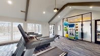 This fitness center is designed to deliver a comprehensive workout experience within a thoughtfully crafted architectural setting. The main exercise area features soaring vaulted ceilings accented by bold exposed dark beams, creating a spacious and energizing atmosphere. A dramatic floor-to-ceiling stacked stone accent wall serves as a focal point, complete with a wall-mounted flat-screen television, adding both style and function to the cardio zone. The space is outfitted with multiple treadmills, elliptical machines, stationary bikes, and adjustable weight benches, providing a well-rounded selection of equipment for both cardio and strength training. A dedicated functional training area houses stability balls, resistance bands, medicine balls, and yoga mats, organized neatly on a multi-tiered rack behind glass-paneled room dividers. Plush patterned carpet tiles in muted blue and gray tones cover the floors throughout. An entire wall of oversized windows and floor-to-ceiling glass panels in the cardio section invites abundant natural light and offers open views of the surrounding landscape and outdoors. Recessed lighting and a pendant ceiling fan ensure the space remains well-lit and comfortable at all hours. The overall ambiance is modern, clean, and motivating.
