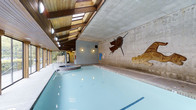 This single-floor indoor aquatic facility offers a thoughtfully designed environment that blends functional amenities with artistic character. The centerpiece of the space is a generously sized indoor swimming pool, its calm, light-blue water reflecting the warm overhead lighting and natural daylight that filters in through a series of skylights set into an exposed wood-beam ceiling. The architectural design features a contrast between the rustic warmth of the timber ceiling structure and the raw, industrial texture of the concrete walls, creating a unique and visually engaging atmosphere. Adjacent to the pool sits a circular hot tub, providing a dedicated space for relaxation. One of the most distinctive features of the facility is the large-scale mural painted directly on the concrete wall, depicting bold Indigenous-inspired artwork with rich earthy tones of brown and gold. Floor-to-ceiling windows along one side of the space invite natural light and offer views of the surrounding greenery, enhancing the overall sense of openness. Wall-mounted lighting fixtures supplement the natural illumination, ensuring the space remains well-lit throughout. Additional service areas and changing facilities are visible in the background, supporting the full functionality of this aquatic center.