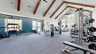 Located at 1485 Fouraker Road in Jacksonville, Florida, this fitness center is a standout amenity space defined by its striking architectural design and comprehensive workout offerings. The single-floor facility is generously sized, providing ample room for a full range of fitness activities without feeling crowded.

The most notable architectural feature is the dramatic vaulted ceiling adorned with rich, exposed wood beams that run the length of the space, lending warmth and character to the otherwise clean, modern interior. Large ceiling fans are mounted along the ridge, ensuring comfort throughout. The crisp white walls are punctuated by a bold teal accent wall, adding a vibrant, energetic atmosphere to the room.

Natural light floods the interior through numerous windows positioned along multiple walls, offering pleasant outdoor views of the surrounding community. The space is finished with durable, patterned carpet flooring suited for a high-traffic fitness environment.

Equipment is thoughtfully arranged throughout the floor, including Matrix-brand treadmills, stationary bikes, a cable and weight machine system, a functional training rig with TRX suspension straps and resistance bands, a rowing machine, weight benches with free weights, and a rack of kettlebells and medicine balls. Wall-mounted mirrors expand the visual depth of the space, while a mounted flat-screen TV provides entertainment for cardio users. The result is a bright, open, and thoroughly functional fitness center.