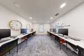 Located at 308 Scott St SW in Atlanta, Georgia, this single-room computer lab office is thoughtfully designed to support a productive and tech-forward work environment. The space is organized around two parallel rows of white marble-top workstations running the length of the room, each equipped with desktop computers and paired with sleek black and orange chairs that add a pop of color to the neutral palette.

The room's white walls are clean and bright, illuminated by recessed ceiling lighting that ensures consistent, glare-free coverage throughout. A standout design feature is the bold navy blue circuit board mural adorning one wall, reinforcing the technology-focused purpose of the space. Complementing this, two large decorative clocks — one with a Roman numeral face and one with a minimalist gold frame — are mounted on the opposite wall, adding character and visual interest.

A centrally positioned window with white blinds allows natural light to filter in, enhancing the open and airy feel of the room. Patterned gray carpet tiles cover the floor, adding texture while maintaining a professional aesthetic. Near the entry, a door with flanking windows provides transparency into adjacent spaces, and a printer station is conveniently accessible. The overall ambiance is clean, modern, and purpose-built for collaborative or individual computer-based work.