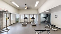 This fitness center offers a well-rounded workout environment designed to support a variety of exercise routines. The space is characterized by soaring ceilings that create an open, airy atmosphere, complemented by a ceiling fan that promotes air circulation throughout. Recessed and panel lighting, combined with large windows along the perimeter, flood the room with natural light and provide views of the surrounding outdoor greenery, enhancing the overall workout experience.

The flooring throughout consists of durable rubber gym matting, providing both safety and comfort underfoot. Exposed light brick columns and walls add a clean, modern aesthetic that ties the space together. A bank of full-length mirrors along one wall visually expands the room and allows users to monitor their form during exercise.

The cardio section features dual treadmills positioned facing the windows, taking full advantage of the natural light and outdoor views. The strength training area is well-appointed with a cable/pulley machine, a power rack, a stationary bike, an elliptical trainer, adjustable weight benches, dumbbells, kettlebells, and medicine balls. The variety and quantity of equipment make this fitness center suitable for both beginner and advanced users, offering a comprehensive, convenient facility for total-body training.