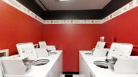 Located at 300 Springboro Lane in Columbus, Ohio, this single-floor laundry facility is designed for functionality and ease of use. The room is finished with a striking red and black color scheme — bold red walls contrast sharply with a black upper trim, creating a visually distinct interior. A decorative border runs along the midline of the walls, adding a subtle detail to the utilitarian space. The ceiling features a textured tile finish with recessed fluorescent lighting that provides bright, even illumination throughout. Multiple top-load washing machines are arranged in two facing rows, maximizing the use of the available floor space and allowing for efficient, simultaneous use by multiple individuals. The machines are white and clearly labeled, contributing to an organized and accessible layout. The flooring features a classic black and white tile pattern, complementing the bold wall colors and adding to the overall clean aesthetic. This laundry facility is straightforward in its purpose, offering a tidy and well-maintained environment equipped to serve the needs of its users efficiently.