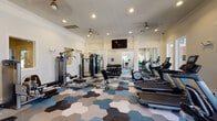 Located at 15270 Voss Rd in Sugar Land, Texas, this well-appointed fitness center offers a comprehensive workout environment within a single-floor layout. The space is defined by its eye-catching hexagonal mosaic carpet in a palette of blue, gray, and white tones, lending a modern and polished aesthetic throughout.

The cardio zone is generously equipped with multiple treadmills, an elliptical, a stationary bike, and a rowing machine, all arranged to face large windows that invite ample natural light and views of the surrounding community. Ceiling fans overhead ensure comfort during workouts.

The strength training area features a full rack of dumbbells spanning a wide weight range, a multi-function cable machine, and several selectorized weight machines, offering a well-rounded resistance training experience. An adjustable workout bench adds further versatility.

Floor-to-ceiling mirrors are strategically positioned along the walls, amplifying the sense of space and supporting proper form during exercise. A wall-mounted flat-screen television is centrally placed for entertainment. Crown molding and clean white walls give the facility a refined, upscale character. Recessed lighting throughout ensures the space is consistently well-lit, creating a bright and motivating atmosphere for fitness.
