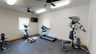 This single-floor fitness room is thoughtfully designed to support a well-rounded workout routine. The space is outfitted with a treadmill, elliptical cross-trainer, and a multi-station strength training machine, providing options for both cardiovascular and resistance training. Dark rubber flooring covers the room, offering durability and traction suitable for active use.

Two ceiling fans are mounted overhead, each fitted with integrated lighting fixtures that, alongside flush fluorescent panels, ensure the room is well-lit throughout. Ventilation is further supported by a ceiling HVAC vent. The neutral, light-colored walls contribute to an open and airy atmosphere, while two windows fitted with white horizontal blinds allow natural light to filter into the space.

A wall-mounted flat-screen television is positioned across from the cardio equipment, enabling users to watch content during workouts. The room's layout is practical and uncluttered, allowing for comfortable movement between machines. Overall, the space provides a clean, functional environment dedicated entirely to fitness and physical well-being.