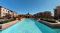 The Main Pool area is a thoughtfully designed outdoor amenity space that offers a resort-style experience. At its centerpiece is a large, sparkling swimming pool with crystal-clear blue water, framed by raised brick water features with tiled spillways that add both visual interest and a soothing ambiance. The pool's elongated layout provides ample room for both leisurely swimming and lounging.

Surrounding the pool is a generously sized sun deck finished in a textured concrete surface, furnished with colorful lounge chairs and shaded by market umbrellas, creating an inviting space for sunbathing. Raised landscaping beds with lush greenery are integrated throughout the deck, adding a natural element to the hardscape design.

A standout feature is the covered outdoor lounge pavilion, constructed with exposed brick walls and open-air archways that frame views of the pool and surrounding buildings. Inside the pavilion, comfortable orange upholstered seating is arranged around a coffee table atop a patterned area rug, alongside a stainless steel built-in grill — ideal for outdoor gatherings. Decorative wall art adds a polished, welcoming touch to the space. The entire area is set against a backdrop of multi-story residential buildings under open skies, enhancing the sense of community and openness.