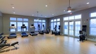 This single-floor fitness center offers a clean, modern environment designed to support an active lifestyle. The space features light hardwood flooring throughout, providing a polished and practical surface well-suited for exercise. The room is generously sized, allowing ample space between cardio equipment including treadmills, ellipticals, and stationary bikes — each outfitted with individual digital displays for an enhanced workout experience. Recessed lighting is complemented by elegant pendant fixtures, ensuring the space is well-lit at all times. Large windows line multiple walls, flooding the interior with natural light and creating an open, airy atmosphere. A set of full-length French doors provides direct access to the outdoors, further enhancing ventilation and connectivity to the surrounding environment. The neutral gray wall tones pair seamlessly with the warm flooring, lending the space a contemporary yet inviting aesthetic. The thoughtful layout and abundance of natural light make this fitness center both functional and comfortable for daily use.