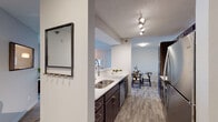 This single-floor residence offers a thoughtfully designed layout that balances functionality and comfort throughout its interconnected spaces. The galley-style kitchen is a standout feature, equipped with stainless steel appliances including a refrigerator, range, microwave, and dishwasher. Dark espresso cabinetry is paired with crisp white countertops, and wood-look plank flooring runs underfoot, complemented by track lighting overhead. A built-in entry nook with a mirror and wall hooks provides a convenient drop zone near the front entrance. The kitchen opens seamlessly into the dining area, which offers easy flow into the living room. The living room features plush carpeting, a large sliding glass door that invites natural light and views of mature trees and green outdoor space, and a wall-mounted AC unit. The primary bedroom is carpeted and well-lit by a generous window, while a second bedroom offers similar comfort and natural light. A flexible den or office space provides additional room suitable for remote work or hobbies. The full bathroom includes dark cabinetry with a white countertop vanity, a shower-tub combination, and wood-look tile flooring, maintaining the cohesive design aesthetic found throughout the home.