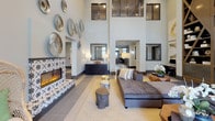 This thoughtfully designed single-floor amenity space makes a striking first impression with a grand, double-height lobby entrance. Floor-to-ceiling windows flood the entryway with natural light, while large-format tile flooring, sheer patterned drapes, and a curated workstation nook with bold abstract artwork set an elevated tone from the moment you arrive.

The central lounge area is anchored by a dramatic patterned tile fireplace flanked by woven accent chairs and a generously sized sectional sofa. Soaring walls are adorned with a gallery of decorative silver disc art and large-scale paintings, contributing to the space's sophisticated, gallery-like ambiance. The double-height ceiling amplifies the sense of openness throughout this area.

Adjacent to the lounge, a dedicated game room provides an inviting entertainment hub, complete with a pool table, foosball table, shuffleboard, and an additional fireplace with matching patterned tile surround. Sheer curtains line the windowed walls, softening the space while allowing natural light to filter in.

The kitchen and dining area features sage-green cabinetry with gold hardware, a marble-top island with bar seating, and a wall-mounted television—ideal for social gatherings. Dark hardwood-style flooring flows throughout the entertainment zones, creating a cohesive and polished aesthetic across the entire space.