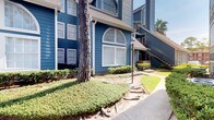 This single-floor unit is part of a multi-story residential complex distinguished by its striking deep blue wood siding, white trim accents, and a prominent arched window that adds architectural character to the exterior facade. Mature trees and neatly trimmed shrubbery line the walkways, contributing to a well-kept, inviting curb appeal.

Inside, the interior presents a contemporary aesthetic grounded in neutral tones and dark concrete-style flooring, which runs consistently throughout the space. The dining area serves as a notable focal point, centered around a round green marble pedestal table paired with woven rattan chairs atop a textured area rug. A large, framed abstract artwork in muted tones adorns the white wall nearby, reinforcing the curated, artistic sensibility of the space.

A sliding barn door with black hardware adds an industrial-modern touch, while a set of vintage-style paneled doors with distressed detailing opens into an adjacent room, introducing eclectic character. Recessed lighting and ceiling-mounted HVAC vents ensure functional comfort throughout. The clean white walls and minimal ornamentation create a calm, gallery-like ambiance within this thoughtfully appointed space.