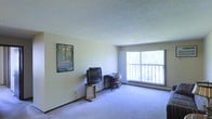 This single-floor space features a generously sized main living area finished with wall-to-wall carpeting in a neutral tone, contributing to a quiet and comfortable atmosphere. The room is characterized by its textured popcorn ceiling, which adds a classic, understated detail overhead. Soft, neutral-painted walls provide a clean backdrop throughout the space.

A prominent sliding glass door dominates one wall, allowing abundant natural light to fill the interior while providing direct access to an outdoor balcony. The balcony, enclosed by a simple railing, offers views of surrounding greenery, enhancing the connection to the outdoors.

The layout accommodates a versatile arrangement, currently configured to function as both a living and workspace area. A dedicated corner near the sliding door serves well as a study or office nook, benefiting from the natural light provided by the large window. The remaining floor space is open and adaptable, suitable for various furniture arrangements.

Overall, the space presents a functional and straightforward design, with its standout features being the natural light, outdoor access via the balcony, and the open, flexible floor plan.