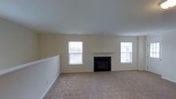 This single-floor space offers a welcoming and well-appointed living area designed with comfort and simplicity in mind. The main living room is generously sized, featuring soft, neutral-toned wall-to-wall carpeting that adds warmth underfoot. A central fireplace serves as the focal point of the room, framed by a clean white mantel that lends a classic, timeless charm to the space. Flanking the fireplace are two double-hung windows dressed with horizontal blinds, allowing for adjustable natural light throughout the day. The smooth, white ceiling and soft cream-colored walls create a bright, airy ambiance that feels open and inviting. The room benefits from a partially open layout, with a low railing visible along one side, suggesting a thoughtful separation of spaces while maintaining visual continuity. Neutral finishes and understated design elements throughout make this space highly versatile and move-in ready, providing an excellent canvas for a variety of interior styles. Overall, the space exudes a calm, livable atmosphere suited for everyday comfort and relaxed living.
