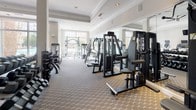 Located at 102 Fallsgrove Blvd in Rockville, Maryland, this fitness center offers a comprehensive and thoughtfully equipped workout facility designed to support a wide range of exercise routines. The space is characterized by its bright, open layout, enhanced by large windows that allow generous natural light to fill the room and provide outdoor views of the surrounding community.

The interior showcases elegant architectural detailing, including crown molding, decorative tray ceilings, white columns, and recessed lighting, giving the gym a polished and upscale ambiance. Ceiling fans are strategically placed throughout to promote air circulation during workouts.

The cardio section features multiple treadmills and elliptical machines positioned near the windows, alongside wall-mounted flat-screen televisions for entertainment. The strength training area is equally well-stocked, with a variety of cable and selectorized machines, a full rack of dumbbells, kettlebells, and medicine balls for functional training. Large mirrors line the walls throughout, enhancing the sense of space and allowing users to monitor their form.

The flooring is covered in a durable, patterned carpet suited for a fitness environment. French-style glass doors provide access to an adjoining area, adding to the facility's open and connected feel. Overall, the space presents a clean, organized, and professionally maintained environment ideal for a complete fitness regimen.