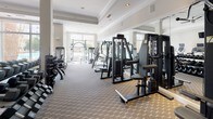 Located at 102 Fallsgrove Blvd in Rockville, Maryland, this fitness center offers a comprehensive and thoughtfully equipped workout facility designed to support a wide range of exercise routines. The space is characterized by its bright, open layout, enhanced by large windows that allow generous natural light to fill the room and provide outdoor views of the surrounding community.

The interior showcases elegant architectural detailing, including crown molding, decorative tray ceilings, white columns, and recessed lighting, giving the gym a polished and upscale ambiance. Ceiling fans are strategically placed throughout to promote air circulation during workouts.

The cardio section features multiple treadmills and elliptical machines positioned near the windows, alongside wall-mounted flat-screen televisions for entertainment. The strength training area is equally well-stocked, with a variety of cable and selectorized machines, a full rack of dumbbells, kettlebells, and medicine balls for functional training. Large mirrors line the walls throughout, enhancing the sense of space and allowing users to monitor their form.

The flooring is covered in a durable, patterned carpet suited for a fitness environment. French-style glass doors provide access to an adjoining area, adding to the facility's open and connected feel. Overall, the space presents a clean, organized, and professionally maintained environment ideal for a complete fitness regimen.