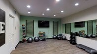 This fitness studio is a thoughtfully designed, single-floor space built to support a wide range of exercise modalities. The room features soft sage-green accent walls paired with neutral tones, creating an energizing yet calming environment conducive to focused training. Recessed ceiling lighting provides bright, even illumination throughout the space.

Along the main wall, two sets of wooden wall-mounted stall bars serve as anchor points for stretching, mobility work, and bodyweight exercises. A flat-screen television is mounted centrally between the stall bars, allowing for guided workout streaming or visual instruction. A large mirror spans a portion of the adjacent wall, enhancing the sense of space and allowing users to monitor their form during exercise.

The studio is stocked with a comprehensive selection of equipment, including a tiered rack of colorful kettlebells in varying weights, a set of medicine balls mounted on a dedicated wall rack, multiple large stability balls, and a neatly organized dumbbell rack. The wood-look flooring offers both durability and comfort underfoot, making the space suitable for dynamic movements, strength training, and flexibility routines.