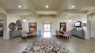 This single-floor community space makes a strong first impression with its open, airy lobby and lounge area. The interior is anchored by a distinctive coffered ceiling with recessed lighting and dual ceiling fans, lending a refined architectural character to the room. Large French doors at the far end of the space flood the interior with natural light while providing a seamless visual connection to the outdoors.

The lounge area is thoughtfully arranged with multiple seating clusters, each featuring red and white checkered upholstered chairs paired with dark bistro-style tables — creating a warm and inviting atmosphere for gathering. A bold, multicolored circular-patterned area rug serves as the centerpiece of the space, adding a lively and modern contrast to the neutral cream and beige wall tones and tile flooring.

Framed artwork on both sides of the room contributes to the welcoming ambiance, while decorative accents such as a large floral vase and a sideboard with a lamp add warmth and personality. The symmetrical layout and generous proportions of the space make it well-suited for socializing and community activities. Overall, the space strikes a comfortable balance between casual and polished, offering a functional yet visually engaging environment.
